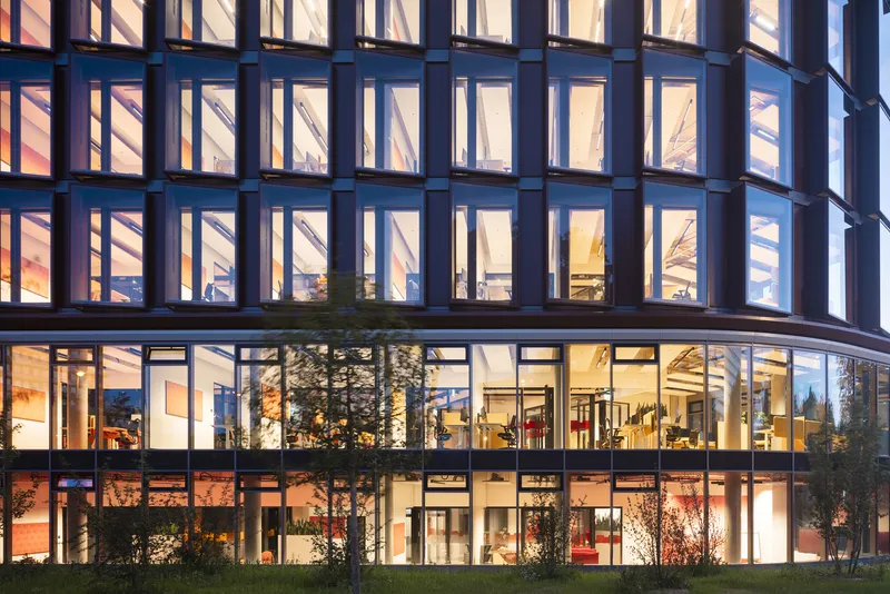 Night view of the illuminated facade showing the grid of windows, ground floor transparency and trees along the base