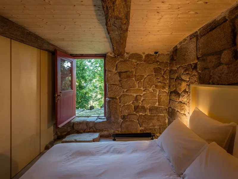 Bedroom corner with exposed granite wall and open red door framing green foliage outside