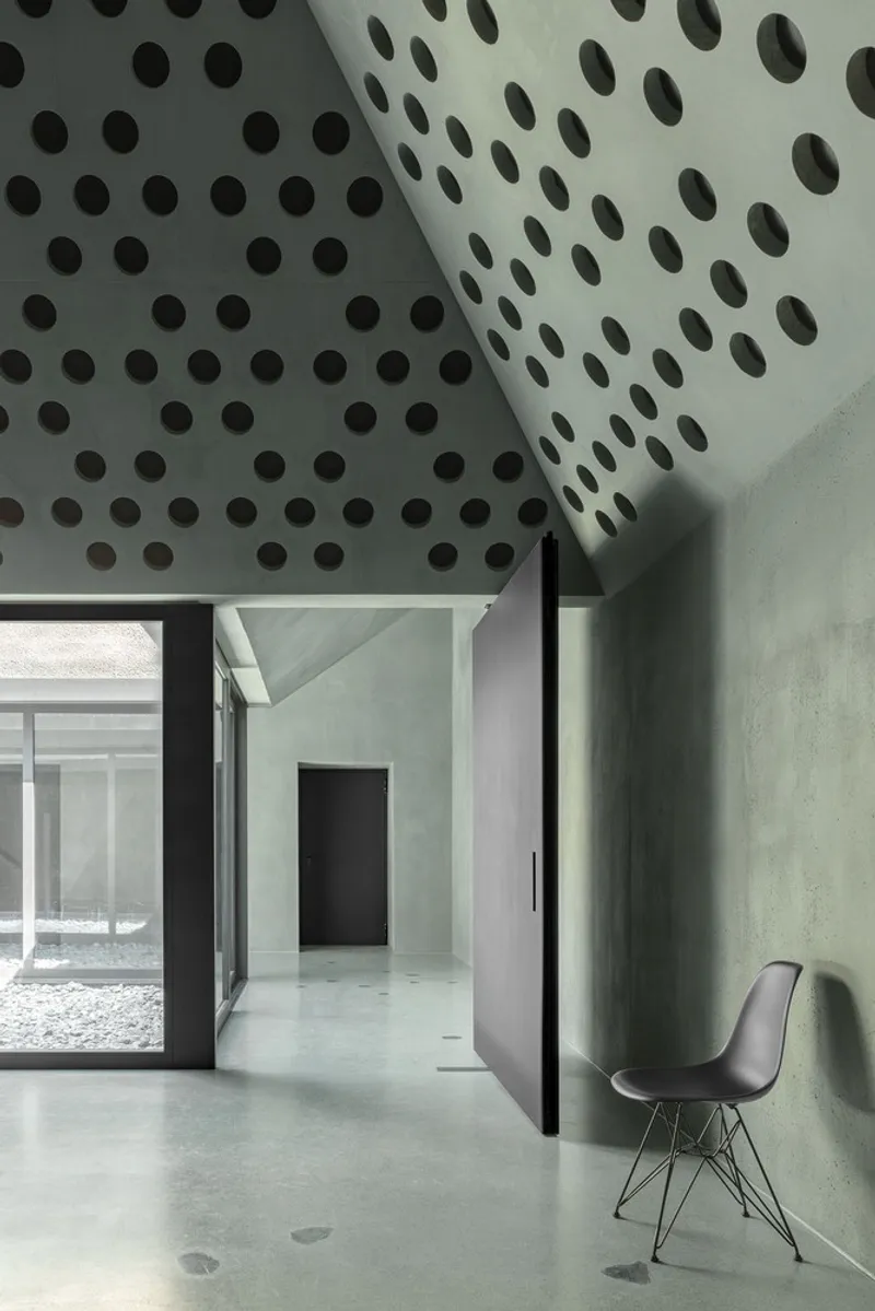 Perforated concrete ceiling with circular openings above a doorway and single chair against green wall