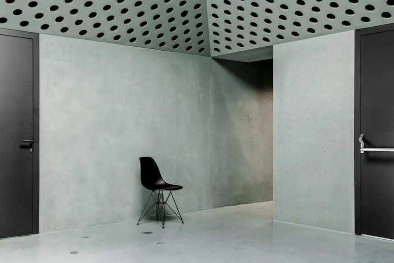 Corner of room with perforated ceiling, polished concrete walls, dark doors, and wire-frame chair