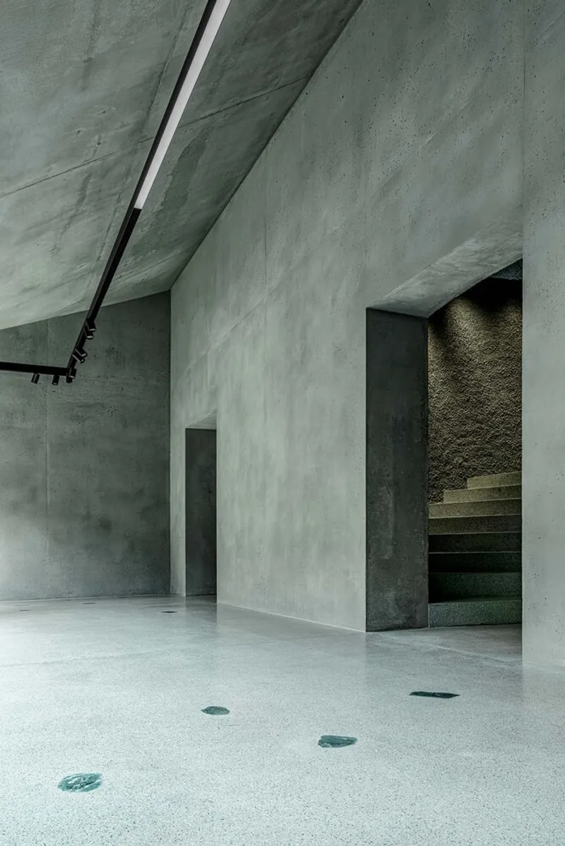 Concrete stairwell opening with exposed treads visible beyond a freestanding wall and angled ceiling