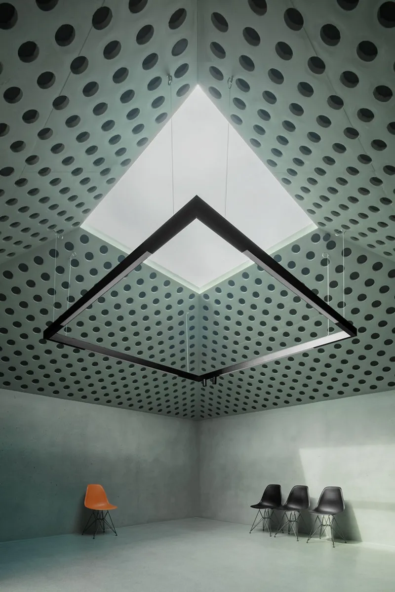 Looking up at pyramidal skylight framed by perforated concrete ceiling with chairs below