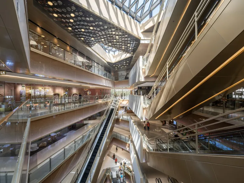 Multi-story atrium with escalators, perforated ceiling screen and daylight filtering through the glass roof above