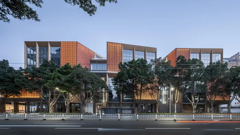 Street view of the multi-level facade with terracotta panels and glass framed by mature trees at dusk