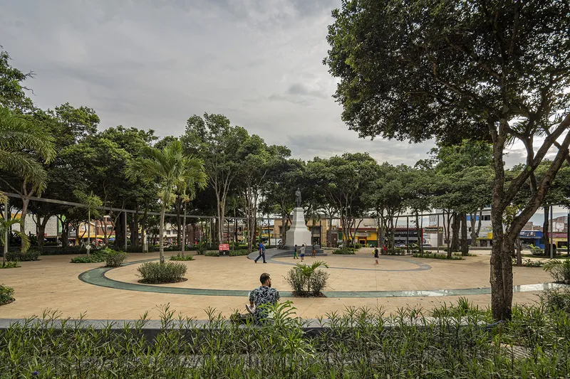 Plaza with sand-colored paving and planted beds beneath a dense canopy of mature trees on an overcast day