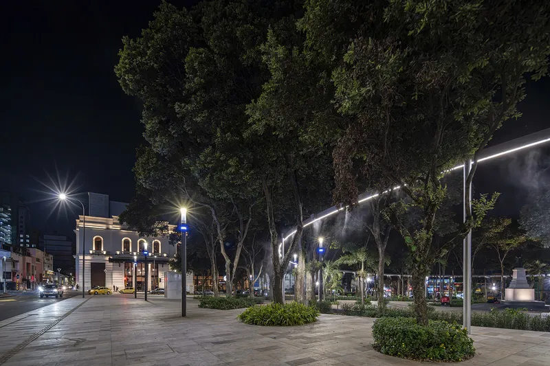 Night view of the plaza edge showing the lit canopy structure and planted beds along the street