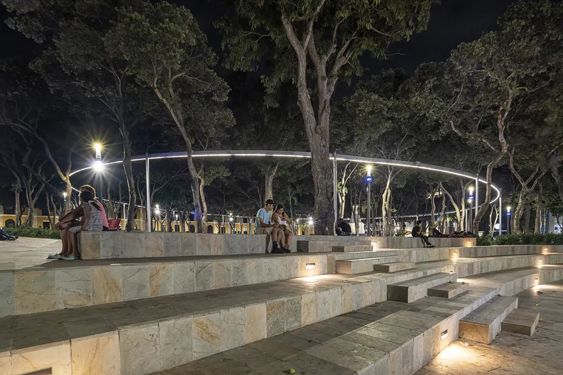Tiered stone seating and steps beneath a curving metal canopy at night with visitors
