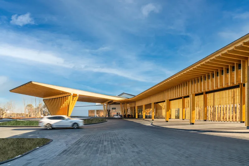 Entrance drive with timber canopy and vertical screen facade under a blue sky with scattered clouds