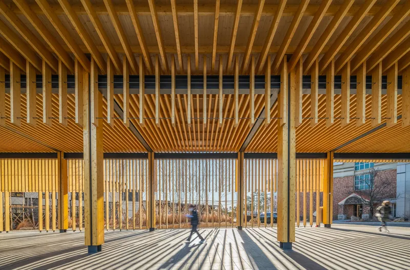 Covered pavilion with vertical timber slats casting shadows on the floor as a figure walks through