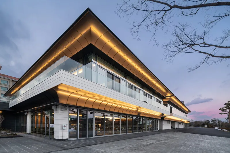 Corner facade with illuminated soffit and glass curtainwall at dusk beneath bare tree branches
