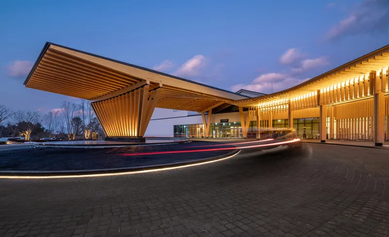 Drop-off area with cantilevered timber canopy and vehicle light trails at dusk