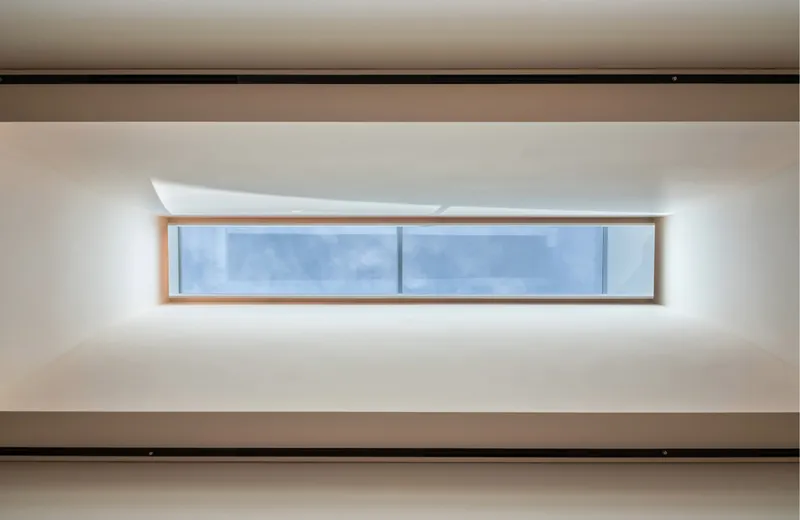 Horizontal slot window with timber frame set into a white ceiling plane beneath a black structural beam