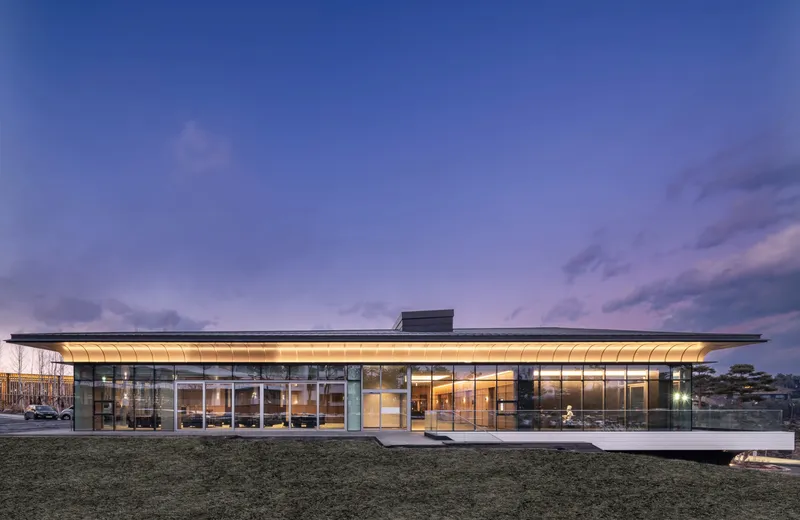 Glass-wrapped pavilion with cantilevered roof and illuminated interior photographed at dusk under a blue sky