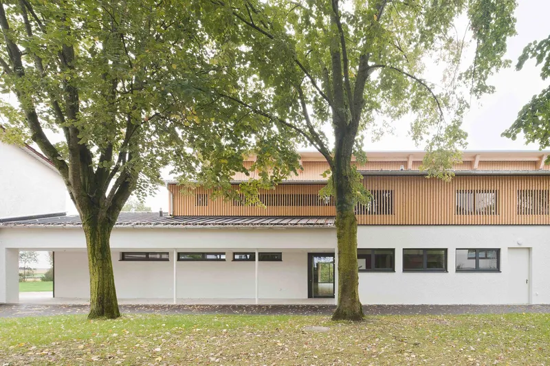 Two-story white rendered facade with timber screens above, framed by mature trees on a lawn
