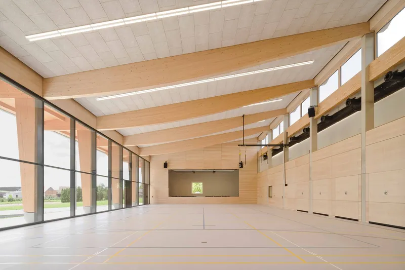 Gymnasium interior with exposed timber beams, floor-to-ceiling glazing and plywood wall panels under linear lighting