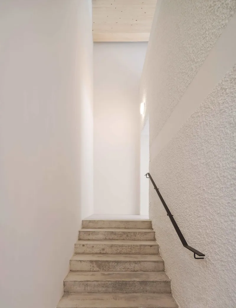 Concrete staircase ascending between white plastered walls with metal handrail and skylight above