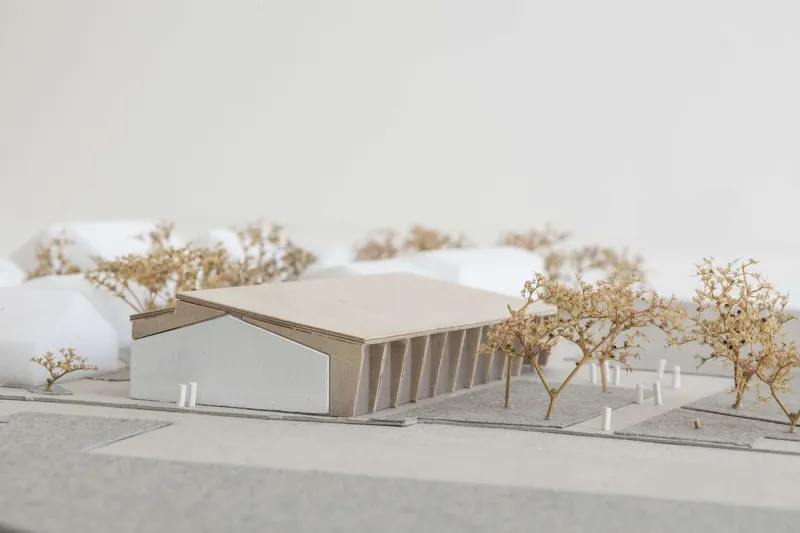 Physical model showing a single gabled structure with corrugated roof among dried trees on a site contour
