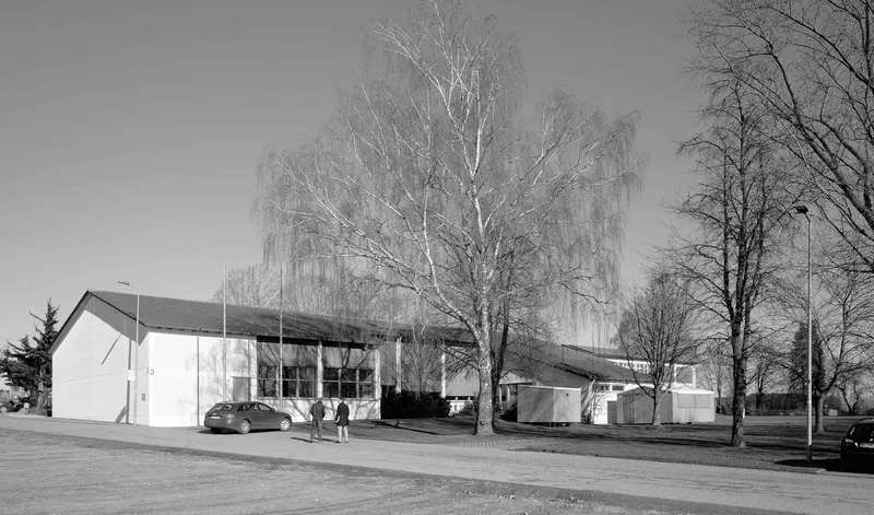 Street view of white-rendered volumes with gabled roof and flat-roofed extension in winter with bare birch trees