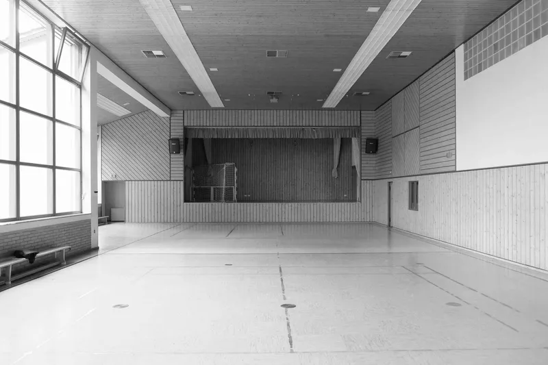 Interior gymnasium hall with timber-paneled walls, acoustic ceiling tiles, stage at far end and gridded steel windows