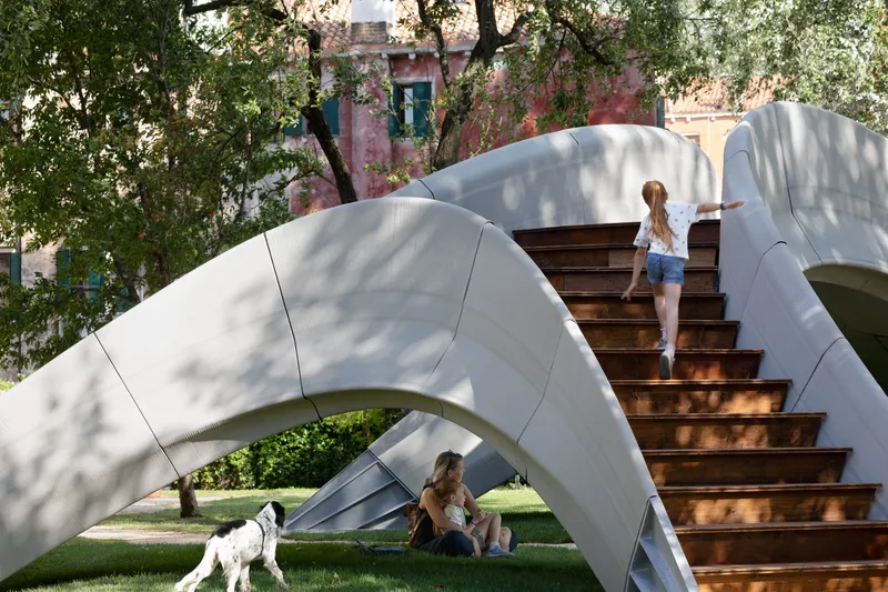 Arched metal shell structure with timber stair and visitors beneath dappled tree shade