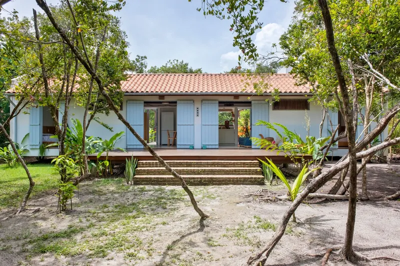 Front facade with pale blue shutters and terracotta roof seen through scattered trees and palms