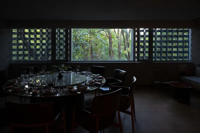 Dining room with perforated concrete screen windows framing a wooded garden in dappled light