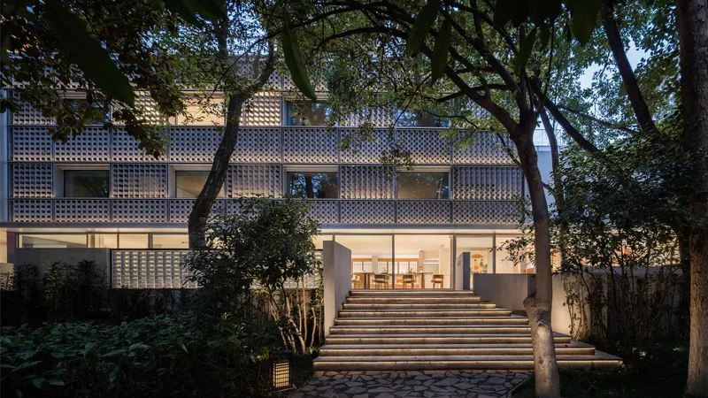Garden courtyard view showing chequered glass block facade, illuminated interiors, and wide entrance steps under evening light