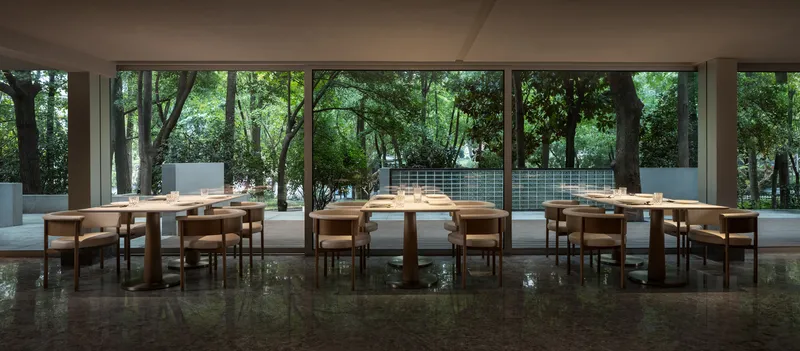 Open dining hall with timber furniture and reflective stone floor opening to courtyard garden through full-height glazing