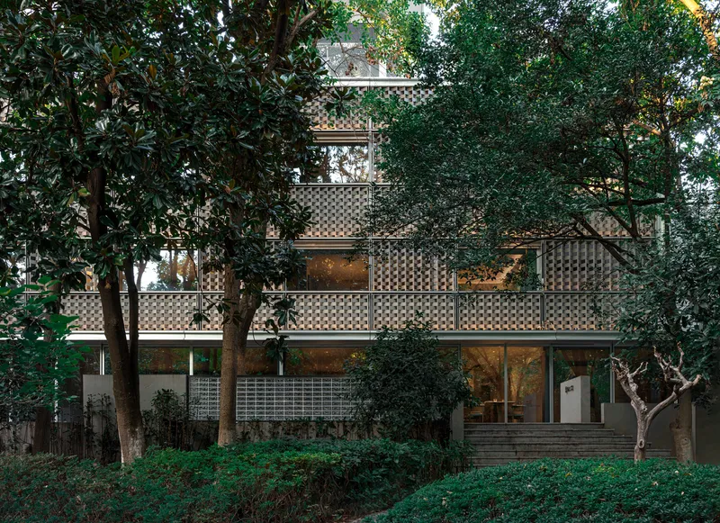 Perforated facade panels framing glazed openings between established trees in the hillside garden