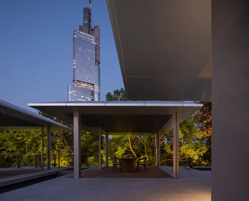 Covered terrace with slender columns overlooking wooded garden and illuminated high-rise tower at twilight