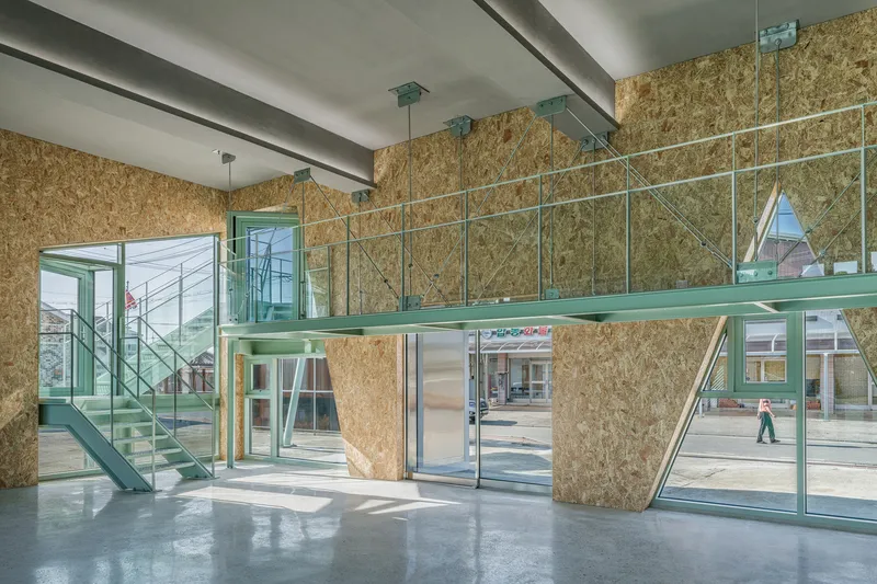 Double-height interior with OSB walls, green steel mezzanine walkway, and glass doors opening to sunlit exterior