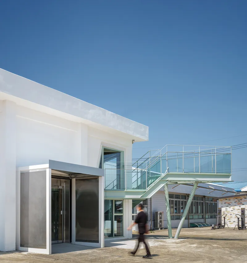 Exterior courtyard with white volumes and a green steel stair bridge connecting rooftop levels as a person walks
