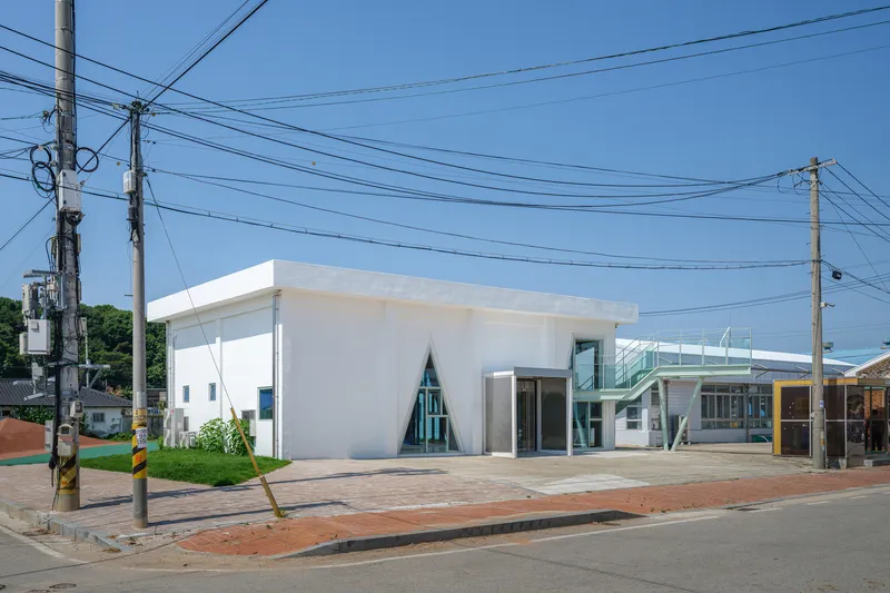 Street view of white concrete volume with triangular window and exposed green steel stair structure under overhead power lines