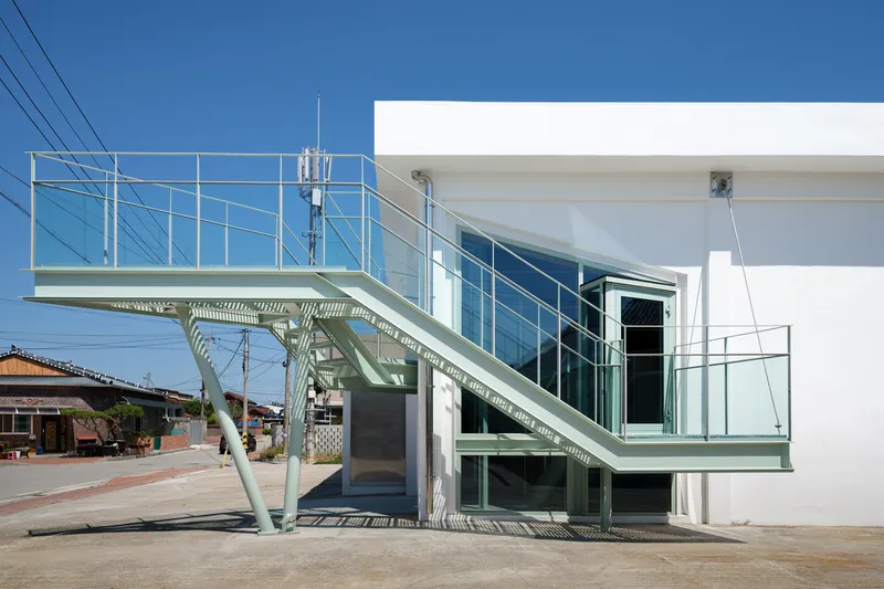 Exterior green steel staircase with splayed tubular supports leading to an upper level entrance on white volume