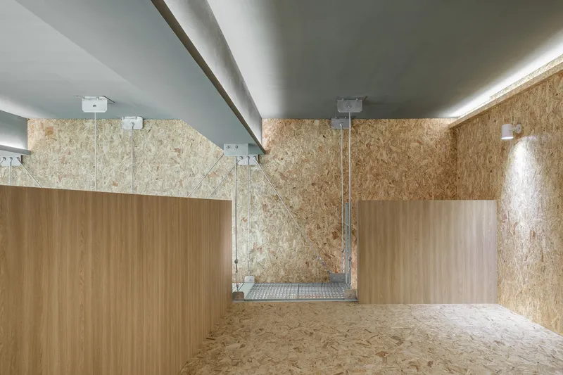 Interior room with OSB panel walls, freestanding timber volumes, and suspended steel mesh platform visible above