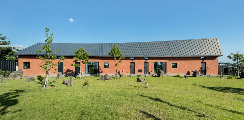Red brick facade with standing seam metal roof surrounded by young trees and lawn in clear daylight