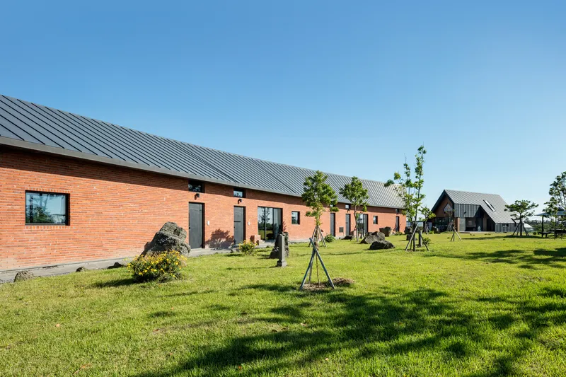 Red brick volumes with metal roofs arranged around a grassy courtyard with staked young trees