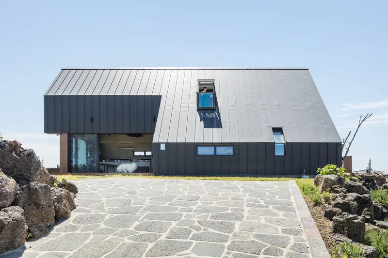 Angular metal-clad volume with steeply pitched roof and stone paving terrace against a blue sky