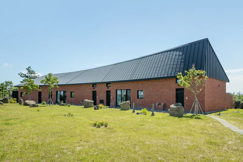 Red brick base with charcoal metal roof volume set in a grass field with scattered boulders