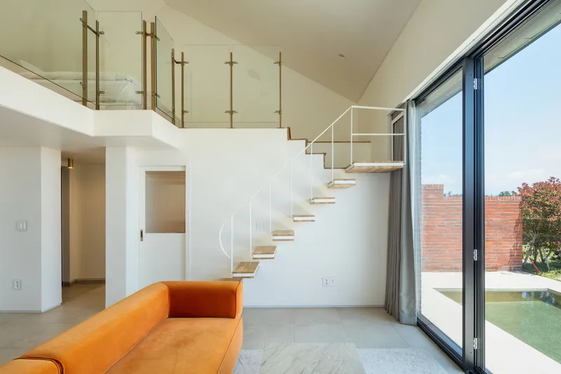 Double-height living room with open-tread timber staircase and floor-to-ceiling glazing overlooking a brick courtyard