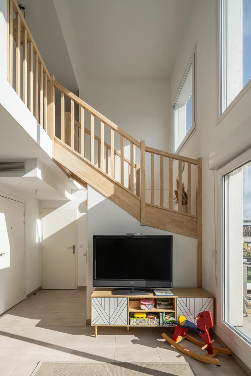 Double-height living space with timber staircase and large windows casting angular shadows across the floor