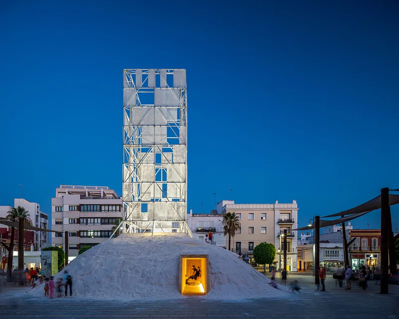 White scaffolding tower above a salt mound with illuminated doorway in a public plaza at dusk