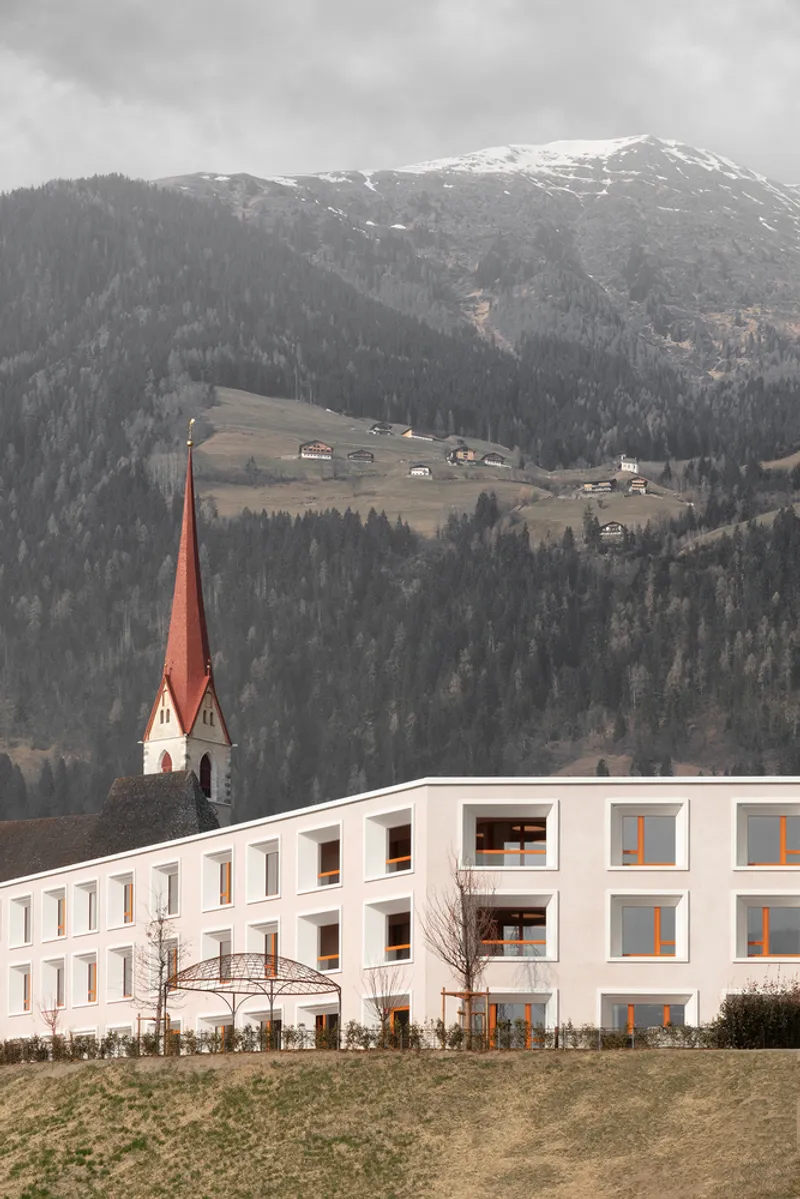 White plaster facade with timber framed windows and balconies against a mountain backdrop with church steeple
