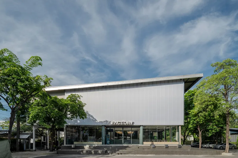 Front facade with sloped metal roof and full-height glazing beneath mature trees on a paved plaza