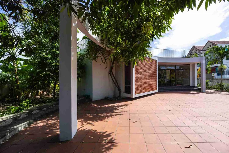 Single-storey volume with perforated terracotta brick screen and red tile paving beneath trees