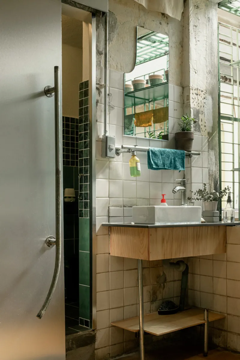 Corner bathroom with white square tiles, timber vanity, and green tile shower beside exposed concrete columns