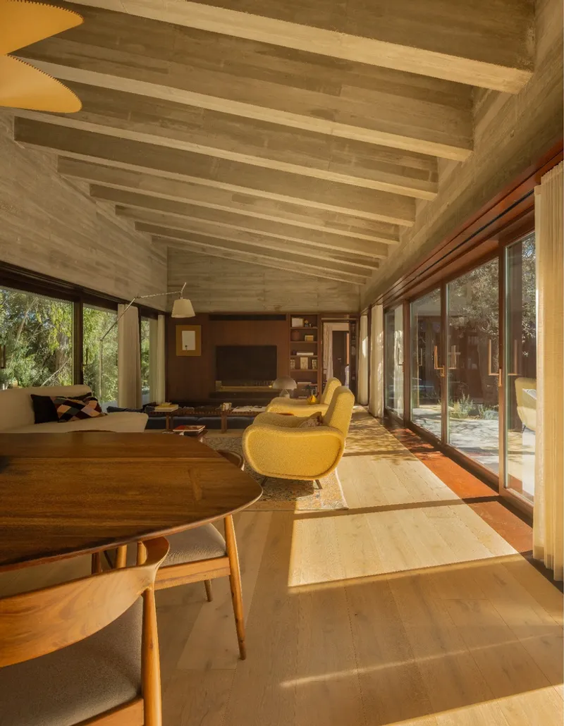 Interior living space with exposed concrete beams and floor-to-ceiling glazing overlooking the courtyard in afternoon light