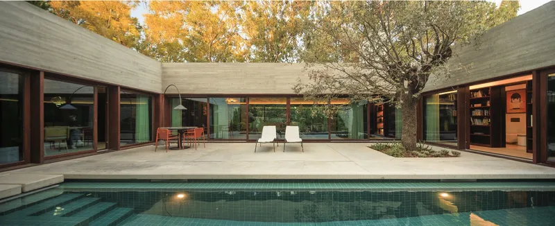 Courtyard pool surrounded by concrete paving and glazed volumes under an olive tree at dusk
