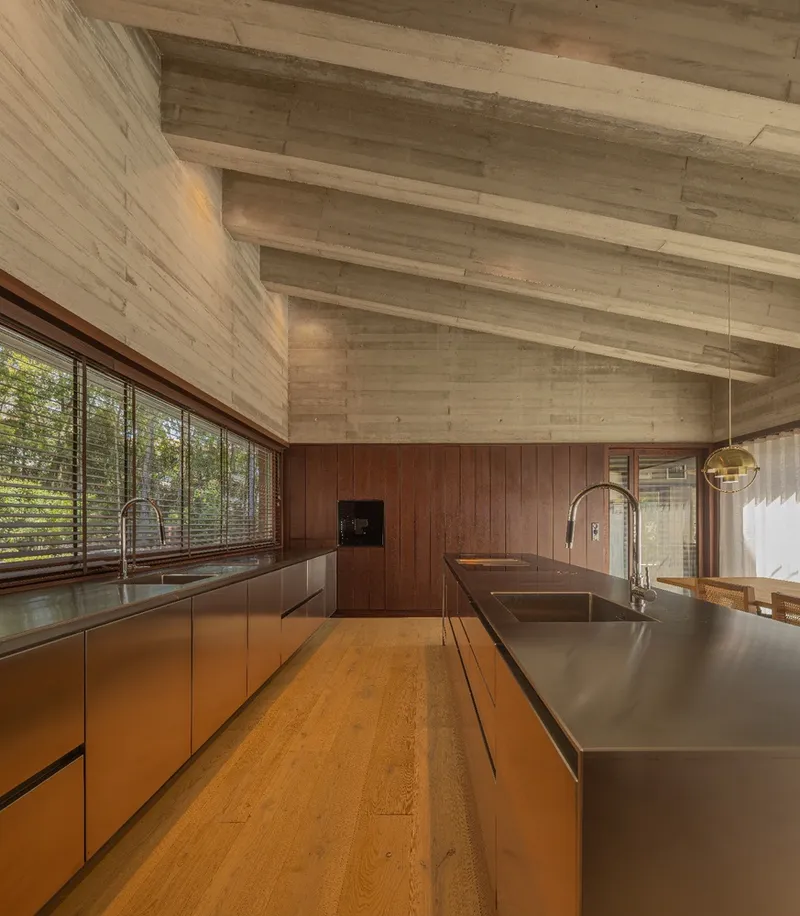 Kitchen with exposed timber ceiling beams and stainless steel island looking toward wood-framed windows into the forest