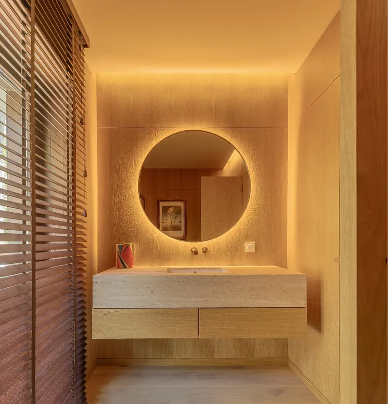 Bathroom vanity in terrazzo with circular backlit mirror and wood louver shutters filtering evening light
